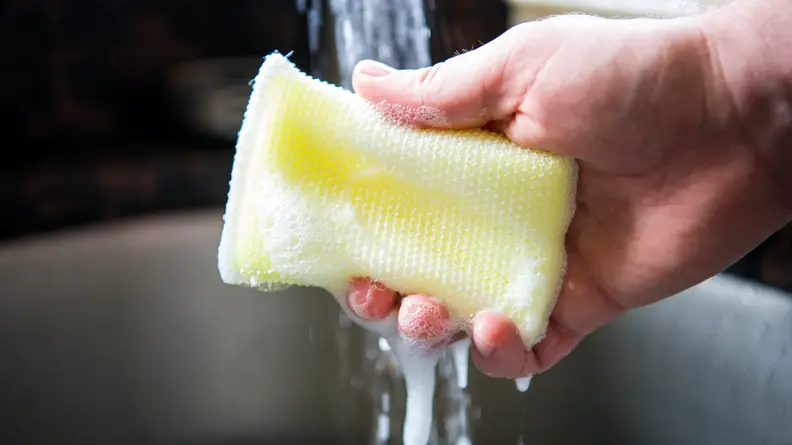 A hand squeezing a Scotch-Brite Dobie sponge in running water