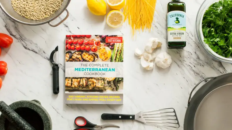 A book sits on a kitchen counter, surrounded by lemons, olive oil, garlic, and various tools. The cover reads, "The Complete Mediterranean Cookbook: 500 Vibrant, Kitchen-Tested Recipes for Living and Eating Well Every Day."