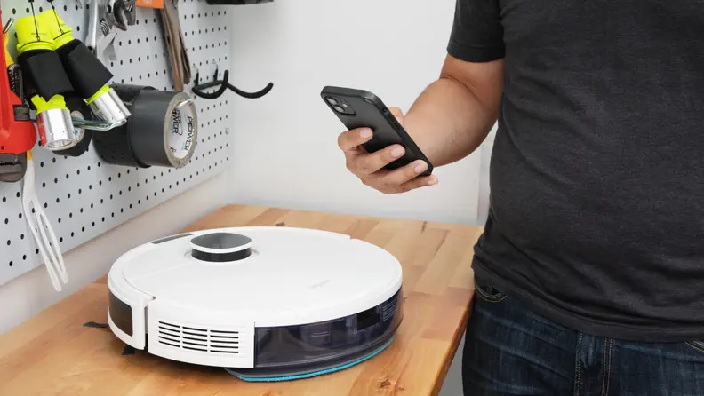 Person hovering smartphone closely to white circular robot vacuum sitting on wooden workbench in front of pegboard with tools.