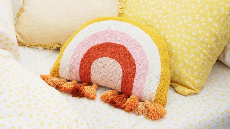 A fringe rainbow throw pillow sits on a bed.