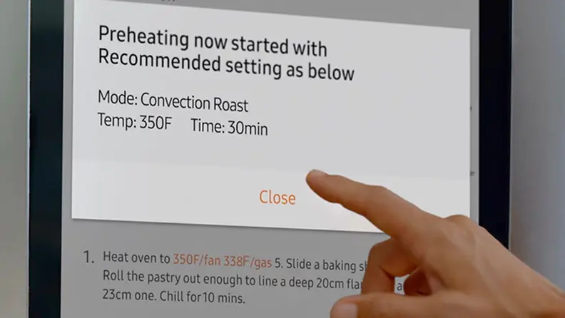 A hand reaches up to a Family Hub display on a Samsung fridge. The extended index finger is about to tap on a dialog box opened up over a recipe, which is asking if the user would like to start preheating the oven.
