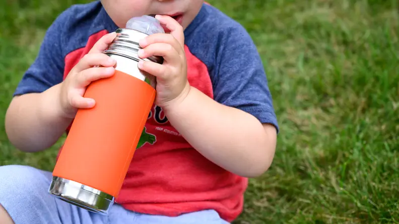 A child sitting on the grass holds a stainless steel Pura Kiki straw cup with an orange sleeve.