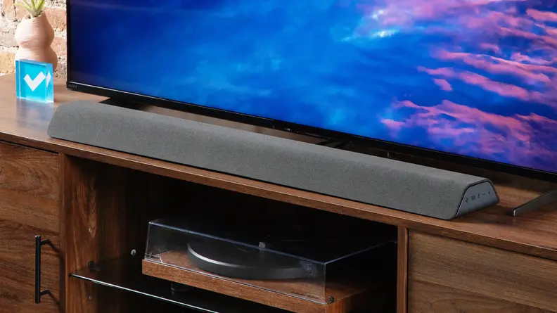 Angled shot of the Vizio M-Series All-in-One M213AD-K8 Dolby Atmos Soundbar resting on an entertainment center in front of a TV.