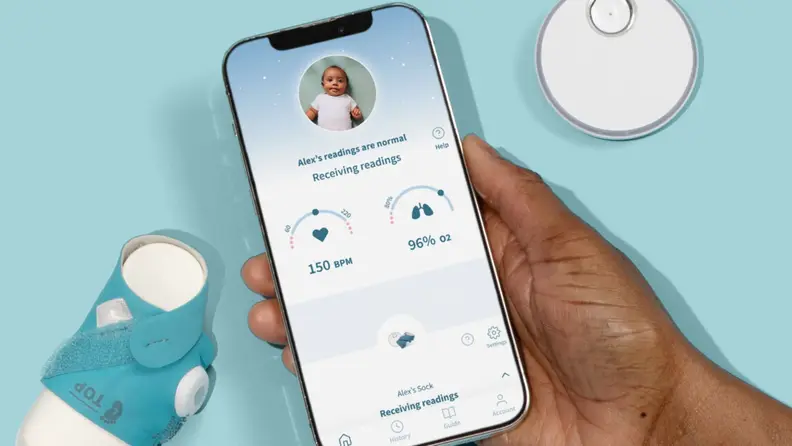 A phone displaying a baby's vitals via the Owlet app. The BabySat sock appears next to it.