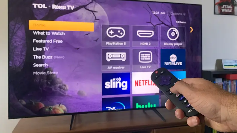 A hand holding a Roku remote pointing at a TCL 5-Series TV displaying the Roku OS home screen.