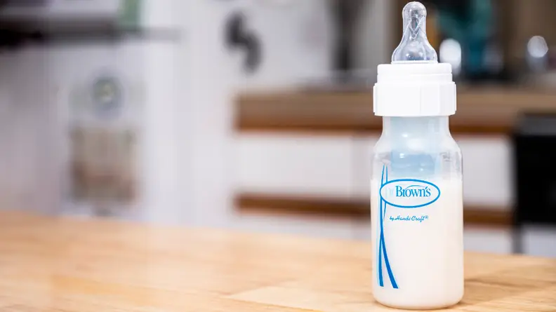 A Dr. Brown's baby bottle on a table with milk or formula inside; the background of a kitchen is blurred out.