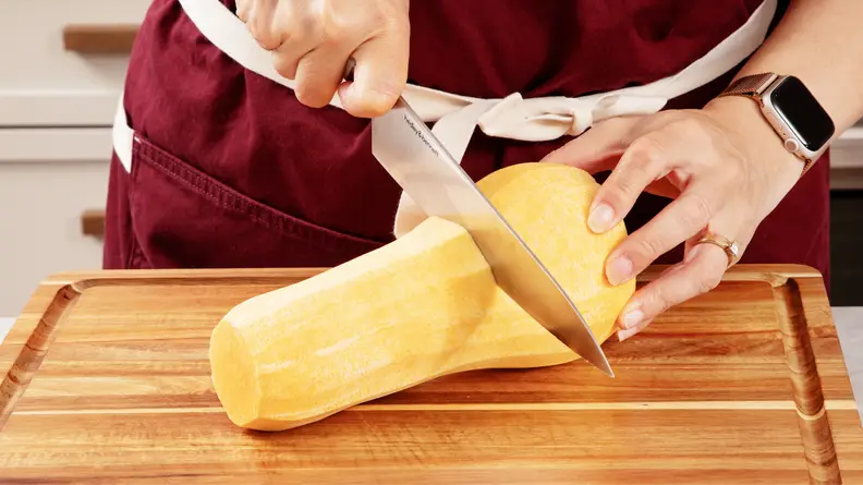 Person using knife to cut butternut squash in half horizontally on top of wooden cutting board.