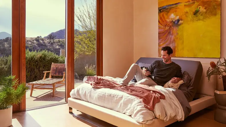 A person reclines on an adjustable bed, smiling at a smartphone while the sun shines in through the windows.