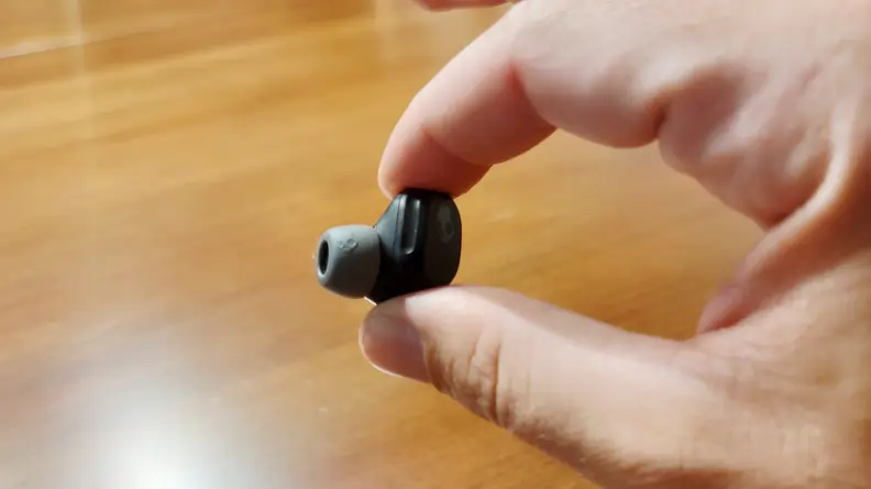 The all-black Skullcandy Mod earbud sits between thumb and finger above a wooden cabinet.
