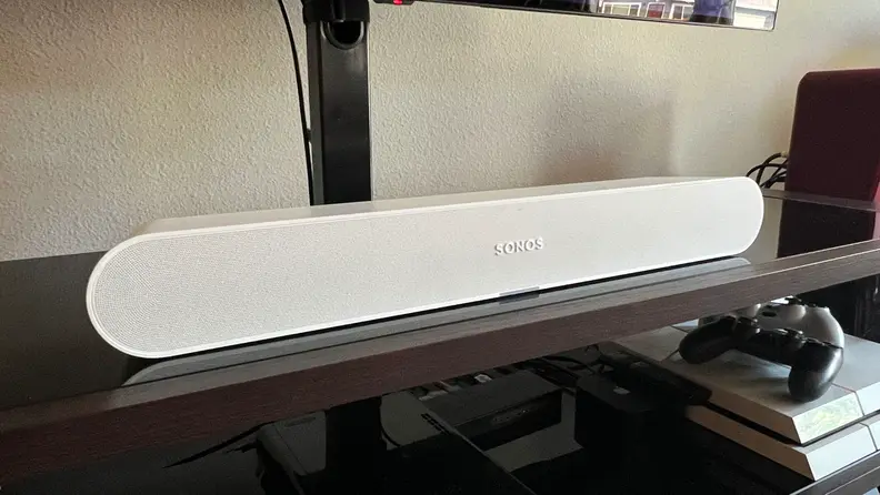 The white Ray soundbar is shown above a PS4 console on a black and brown TV console.