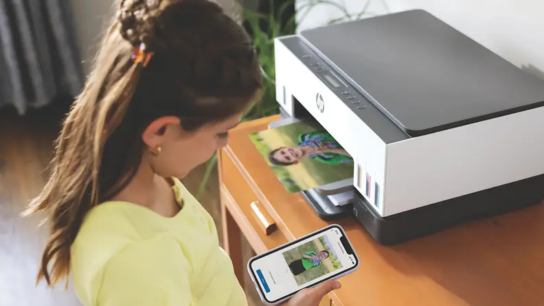 A young girl uses her smartphone to print a photo wirelessly on an HP Smart Tank printer, watching the vibrant image emerge onto the output tray.