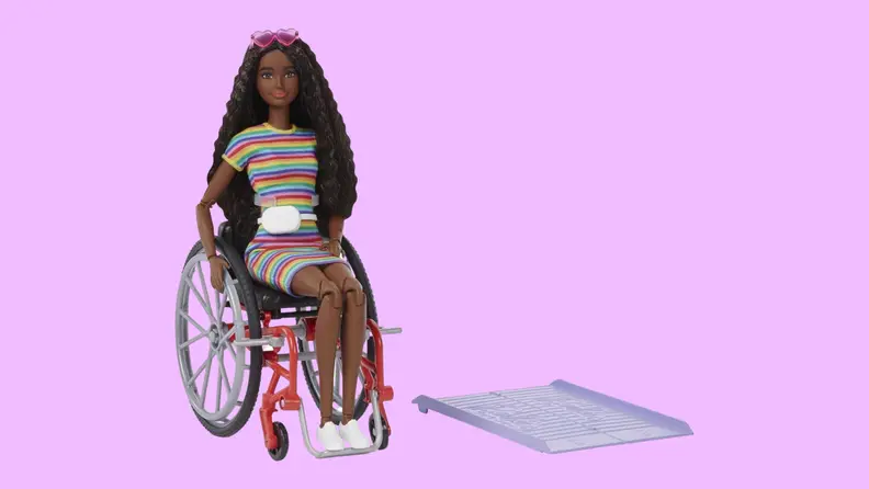 A Barbie in a rainbow-colored dress sits in a wheelchair next to a ramp.