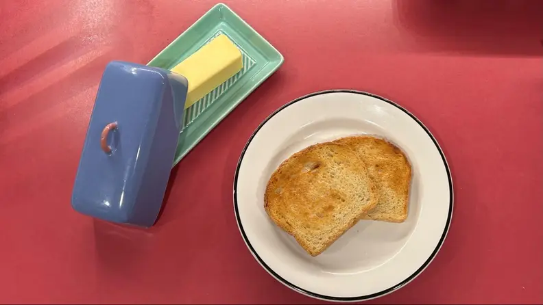 A white plate with a piece of toast on it, next to a blue glass tray with a stick of butter on it.
