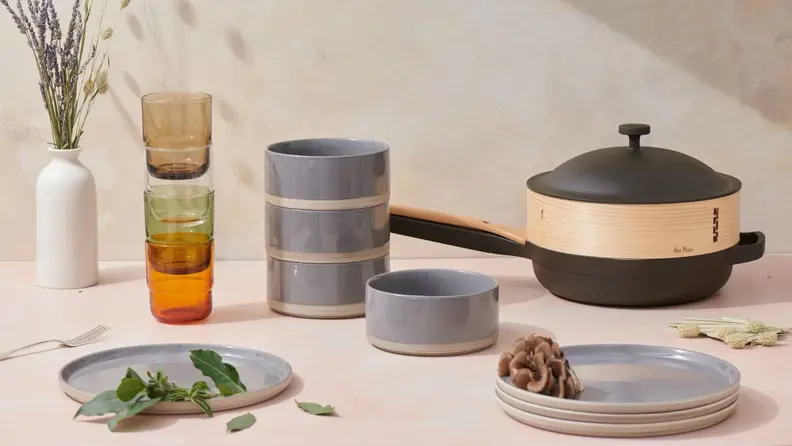 Dark grey plates and bowls arranged artfully on a marble surface next to a pan, stacked glass cups, and a vase filled with flowers.