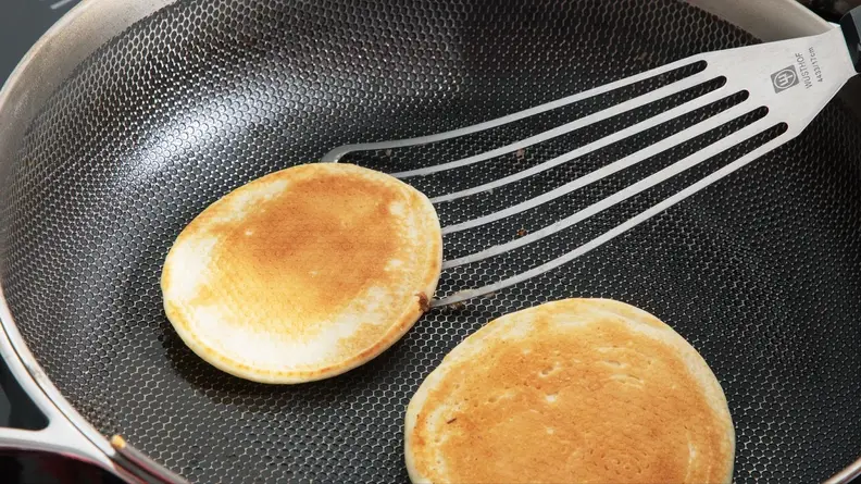 Two golden brown pancakes in the process of being flipped with spatula inside of the Always Pan Pro.
