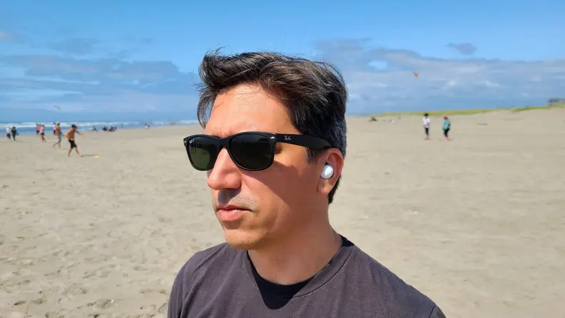 Samsung Galaxy Buds 2 is shown in the ear against a sunny beach, the listener is a man with brown hair wearing sunglasses.