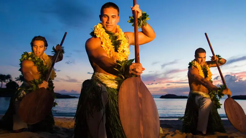 Three Polynesian dancers perform a traditional paddle dance on a beach at sunset