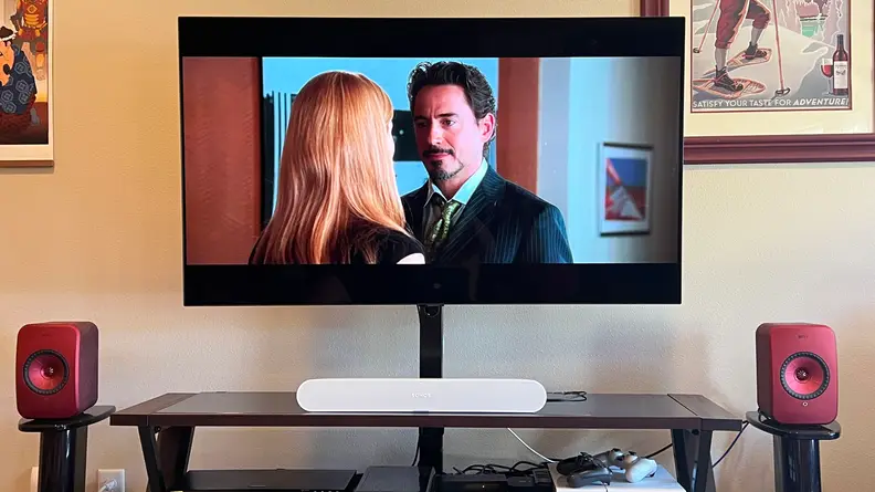 The Sonos Ray sits on a brown TV console between red speakers with Iron Man shown on screen.
