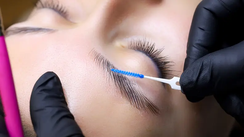 A closeup on a person's eyebrows while a gloved hand runs a spoolie through the hairs.