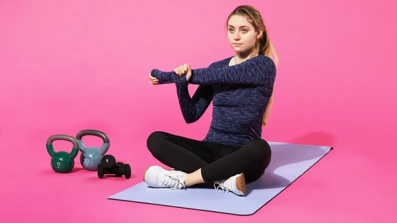 Person sitting cross-legged on Lululemon Workout Mat while stretching left arm across their body next to kettlebells.