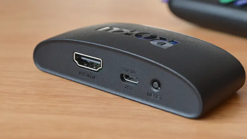 The all-black Roku Express 4K+ sits atop a yellow woodgrain table with its back ports showing, including small power port and an HDMI port.