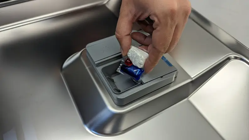 A multicolored dishwasher tablet being placed into a dishwasher detergent dispenser.