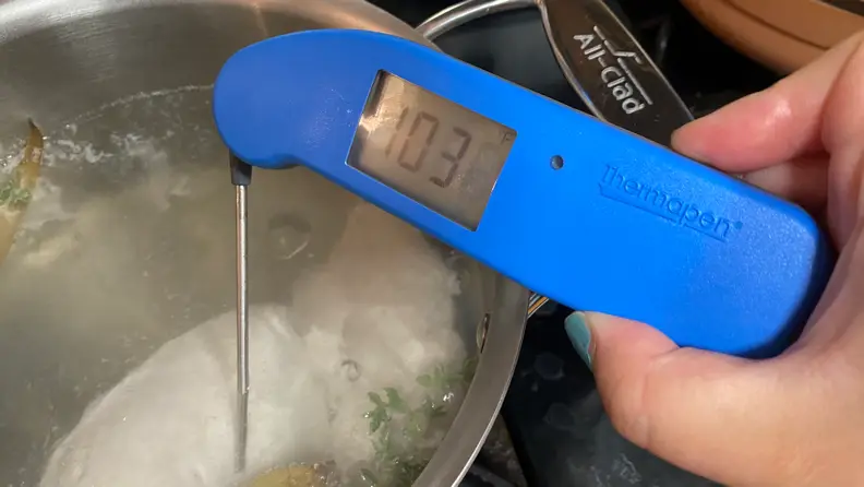 A person is poking a Thermapen ONE digital meat thermometer in Blue into a piece of poached chicken in a large pot.