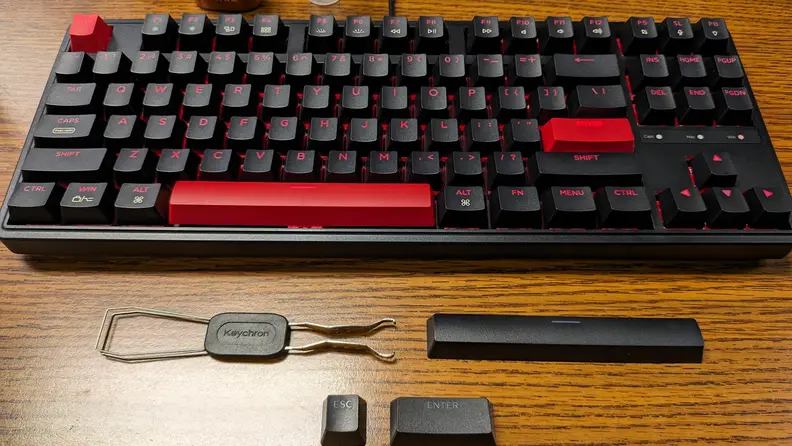 Overhead view of the Keychron C3 Pro laid out on a brown tabletop with its two-in-one keycap and switch remover tool and the ESC, Enter, and spacebar keycaps.