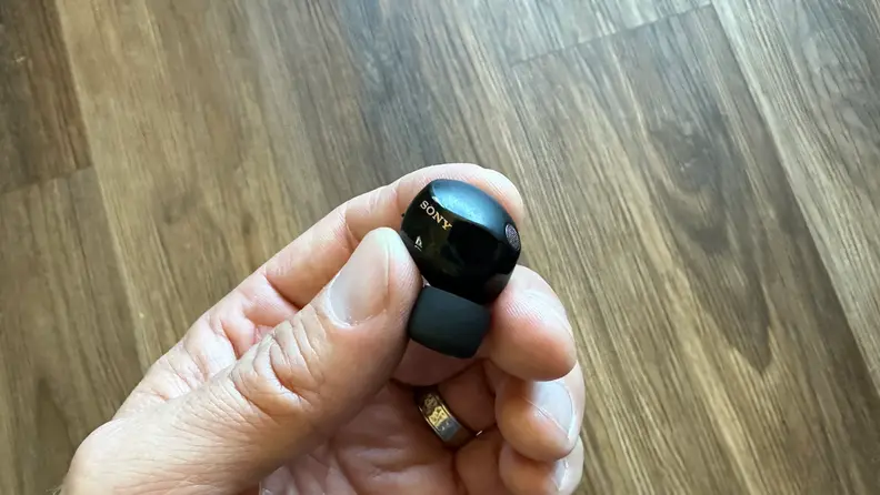 A hand holding one Sony WF-1000XM5 earbud in its fingers.