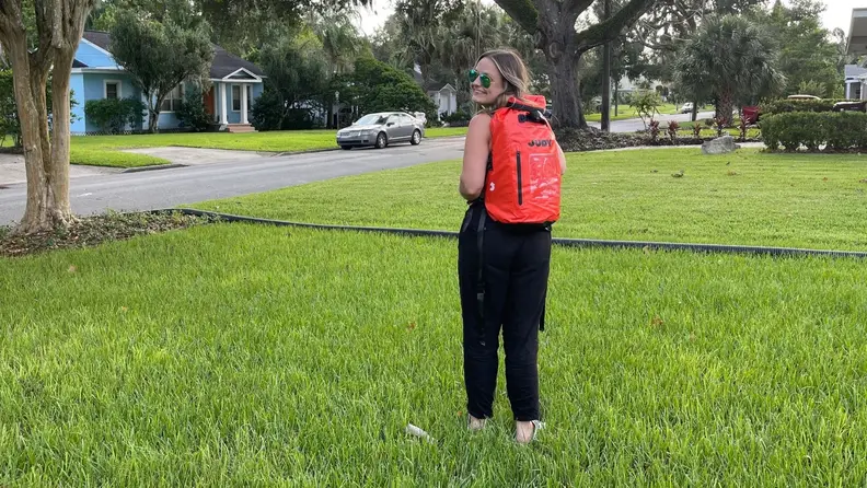 A person stands in the yard wearing an orange Judy backpack.