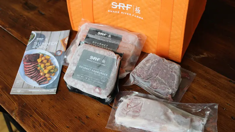 From the left, there's a brochure explaining what Snake River Farms is, a package of ground Wagyu beef, a package of Wagyu hot dogs, a piece of steak, and a piece of pork chop. An orange Snake River Farms bag is in the back.