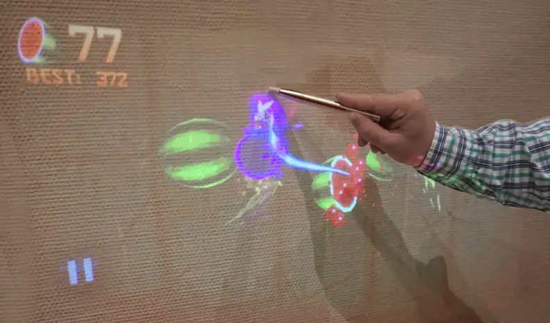 A demonstrator plays Fruit Ninja on a wall in the exhibit hall using the TouchPico.