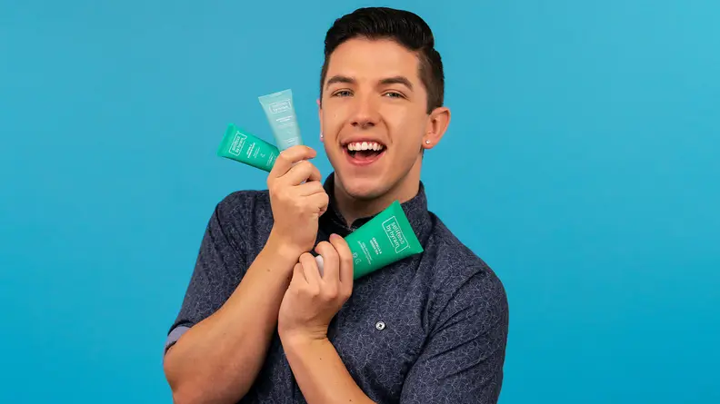 Hyram Yarbro, the founder of Selfless by Hyram, on a blue background holding up three products from his line.
