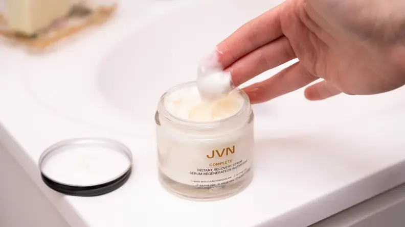 A jar of hair serum with a hand picking up a dollop of the white cream.