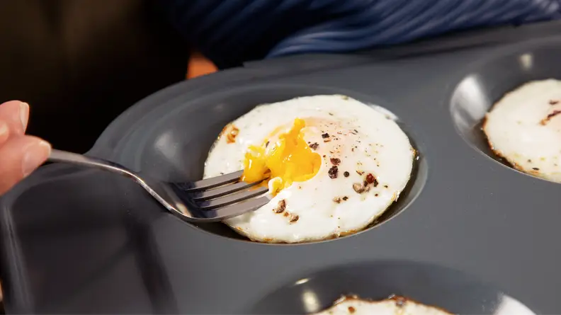 A person using a fork to assess the yolk of an egg that's inside of an egg cooking tray.
