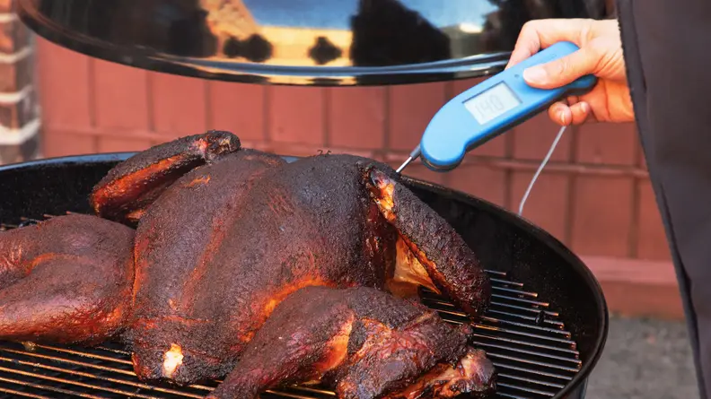 Person using a Thermapen to check the temperature of smoking turkey on a charcoal grill outdoors.