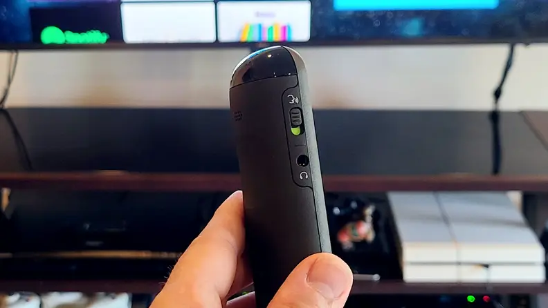 A hand holds the black, rounded remote in front of a TV console with a hands-free switch showing.