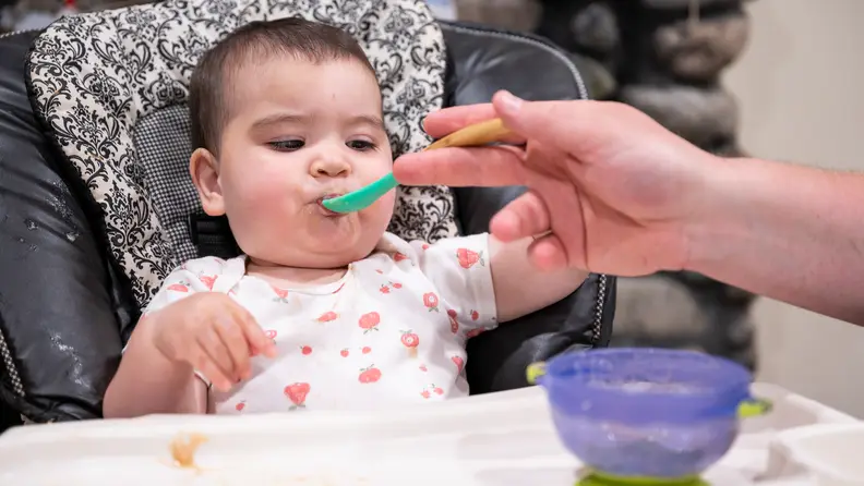 A baby sitting in a high chair being fed by a parent.