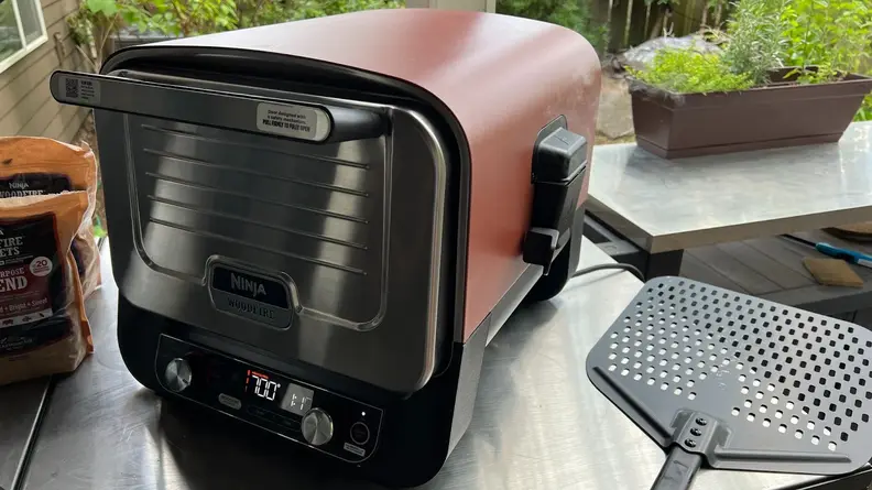 Ninja Woodfire 8-in-1 Outdoor Oven on top of stainless steel countertop outdoors next to bag of pellets and perforated pizza peel.