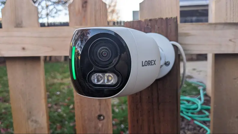The Lorex 4K Dual-Lens Wi-Fi Security Camera displays a green light on the front and hangs outdoors on a fence post.