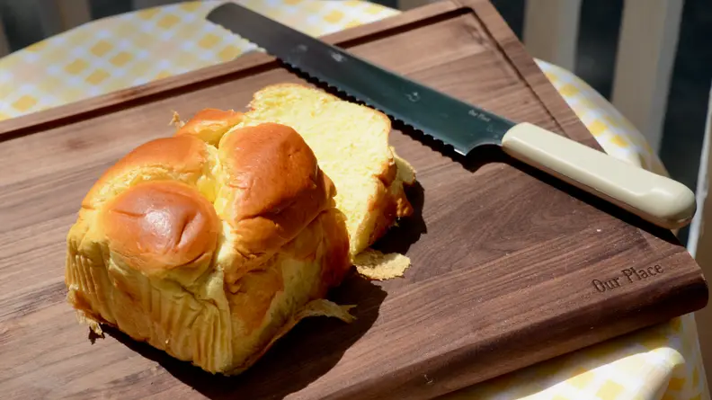 On an Our Place walnut wood cutting board, there's a loaf of brioche bread. Next to it, there's a serrated slice knife.