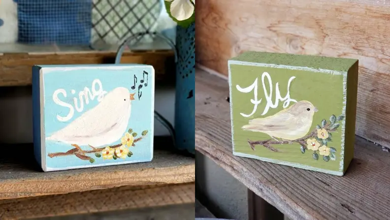 On left, blue wooden garden sign with hand painted white bird on front that reads "Sing."  On right, wooden garden sign with hand painted white bird on front that reads "Fly."