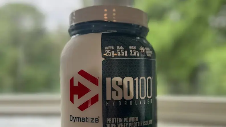 Close-up view of a bottle of Dymatize protein powder in front of a lush green background.