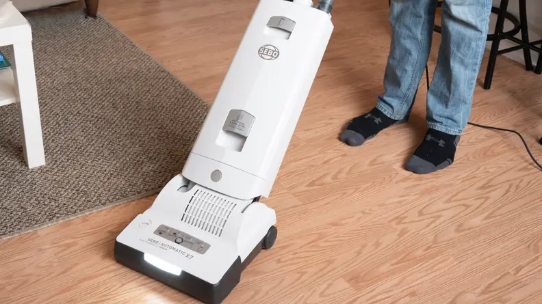 A person in black socks and jeans holds a white upright vacuum on a wood floor