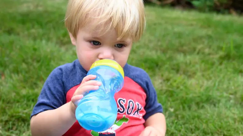 A blonde child in a Red Sox shirt sits on grass and sips through a blue Nuby Clik-it Flex Straw cup.