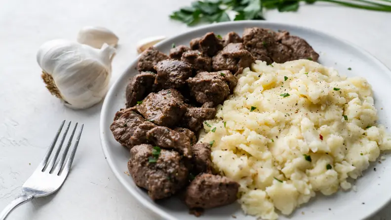White plate with chopped beef and mashed potatoes next to a full clove of garlic