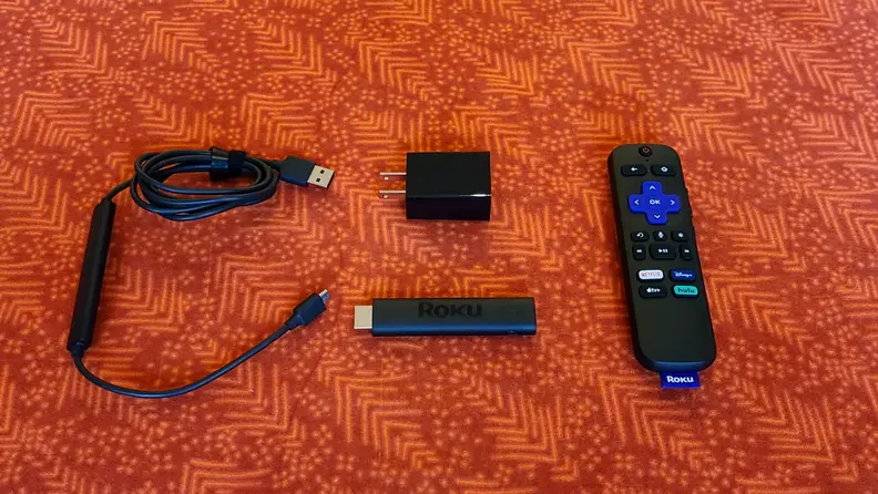 The matte black, candy-bar sized Roku Streaming stick is held with its port showing above a orange-red table with accessories.