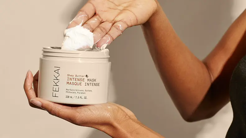 A person holding a gray jar and scooping out a white cream with their other hand.