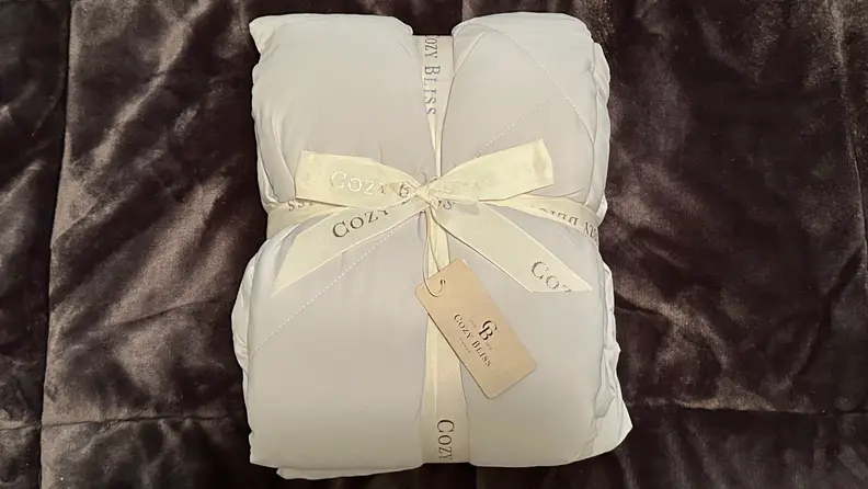 Gray Cozy Bliss comforter folded into a square cushion shape tied together with a white ribbon, laying on a brown fuzzy blanket.