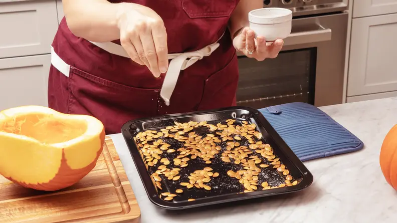 A person holding a white bowl, seasoning the naked pumpkin seeds on the metal sheet.